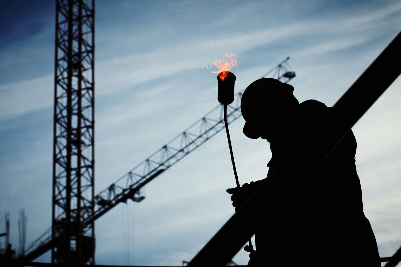 services-03 Silhouette of a construction worker using a blowtorch at a building site against a crane-filled skyline.
